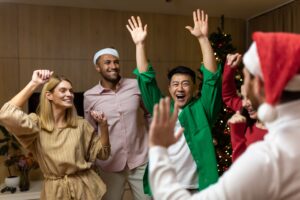 A group of people wearing festive clothes and Santa hats smile and raise their arms in celebration indoors near a decorated Christmas tree.