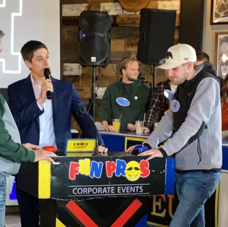 A man in a suit holds a microphone next to another man at a game show-style podium labeled "FUN PROS CORPORATE EVENTS" in a room with several people in the background.
