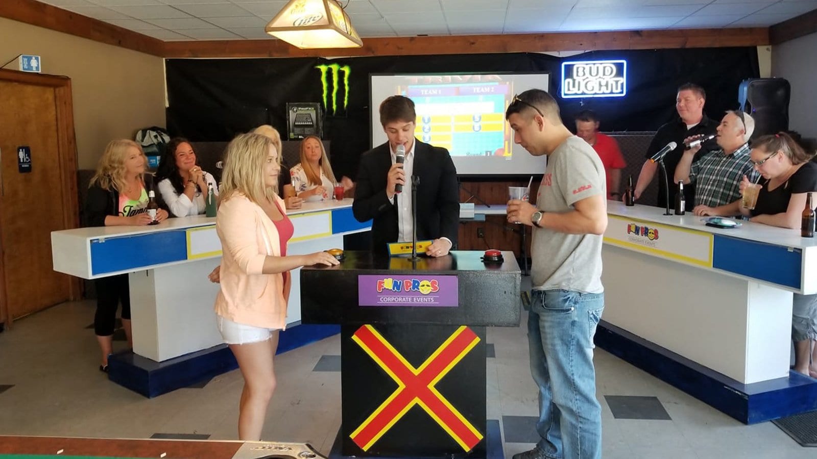 Two contestants stand at a game show podium with a host, while the audience watches in the background. The Chicago Game Show Experience features a Family Feud-style game board displayed on a screen.