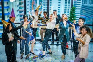 A group of people in party hats celebrate indoors with confetti, drinks, and a small Christmas tree near large windows overlooking a city.